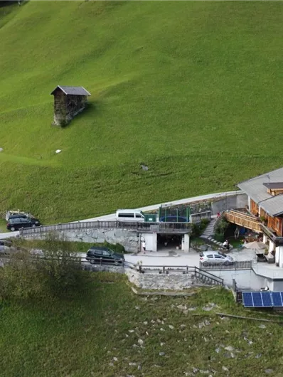 A cozy house on a green meadow, surrounded by mountains. A winding road leads to the building, which is equipped with solar panels.