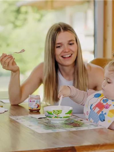 Eine lächelnde Mutter sitzt mit ihrem kleinen Kind am Tisch. Beide genießen ihr Essen in einer gemütlichen Umgebung.