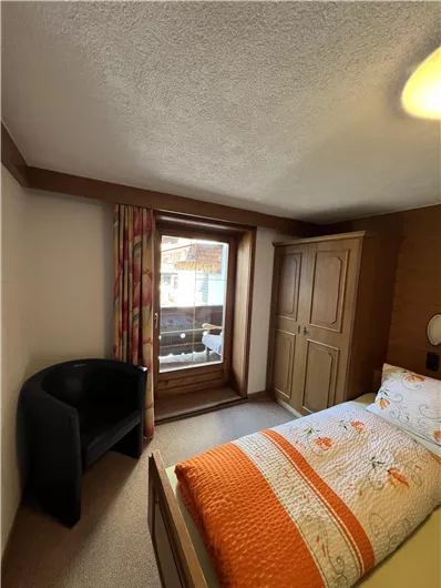 A cozy bedroom with a single bed and a view of the balcony. The room is simply furnished with an armchair and a wardrobe.