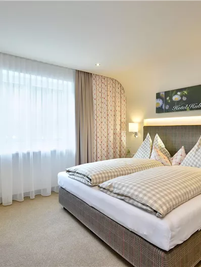 Ein modernes Hotelzimmer mit einem Doppelbett und gemütlichen Kissen. Das Zimmer ist hell und freundlich, mit Tageslicht und eleganten Vorhängen.