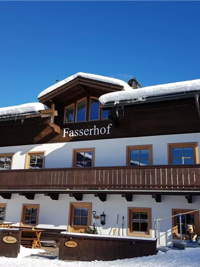 Ein traditionelles Chalet im Schnee mit einem klaren blauen Himmel. Das Gebäude hat einen Balkon und den Namen "Fasserhof" an der Fassade.