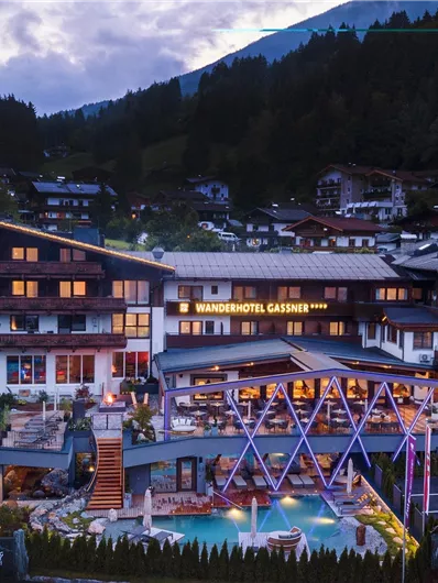 A picturesque hotel in the mountains with a beautiful pool area. The buildings are designed in a traditional style and are surrounded by forest.