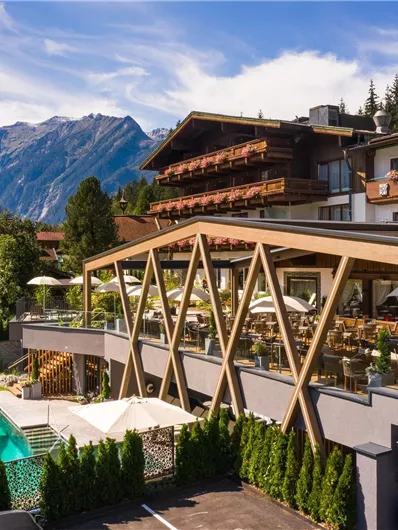 Ein modernes Hotel mit einem einladenden Außenbereich und einem Pool. Im Hintergrund sind die majestätischen Berge und eine grüne Umgebung zu sehen.