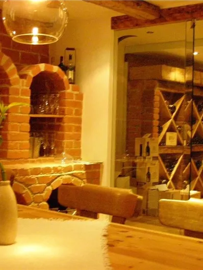 Ein gemütlicher Raum mit roten Backsteinwänden und einem Holztisch. Im Hintergrund befindet sich eine Glastür sowie Regale mit Flaschen.