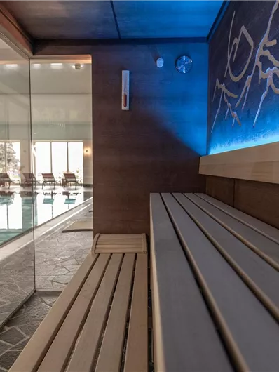 Ein moderner Saunabereich mit Holzbank und blauer Beleuchtung. Im Hintergrund ist ein Pool mit Liegen sichtbar.