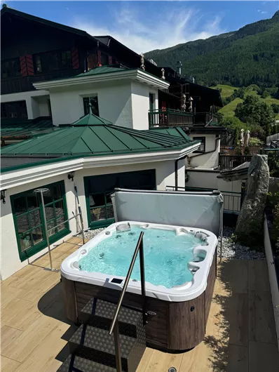 Ein moderner Whirlpool auf einer Terrasse mit Blick auf die Berge. Die Umgebung ist geprägt von grünen Wiesen und einer idyllischen Architektur.