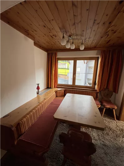 A cozy room with wooden walls and a large table. Orange curtains can be seen in front of the window.