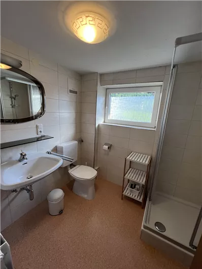 A modern bathroom with a shower, sink, and toilet. It has a large mirror and a bright window view.