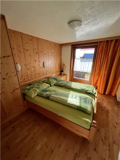 A cozy bedroom with a double bed and wooden paneling. Large windows let in plenty of daylight and offer a view outside.