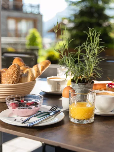 An outdoor breakfast table with fresh rolls, yogurt, fruit, and drinks. The surroundings are green and sunny, ideal for a relaxed start to the day.