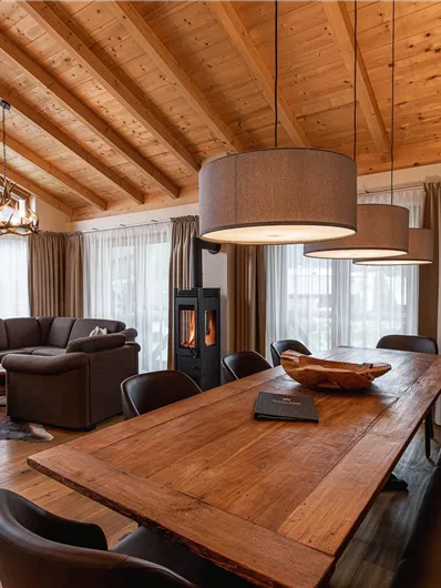 A modern living room with a wooden ceiling and large windows. The dining area features an elegant wooden table and comfortable chairs.