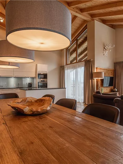 A modern living room with wooden floors and large windows. In the foreground, there is a wooden dining table, surrounded by elegant chairs and stylish lamps.