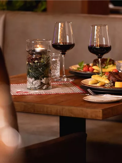 A stylishly set table in a restaurant with delicious food and red wine. In the background, there are candles and decorative plants.