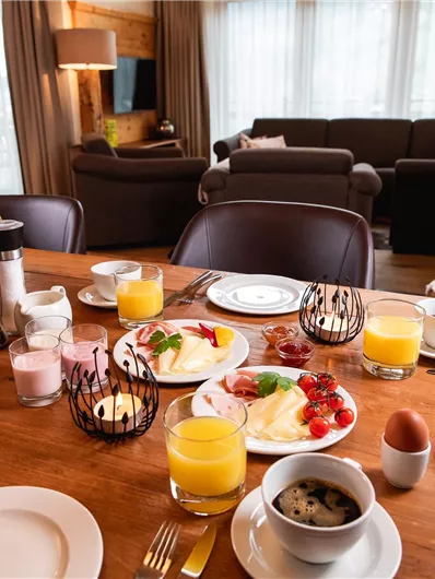 A lavish breakfast table with a selection of rolls, fruit, and beverages. In the background, a cozy living room with a fireplace is visible.