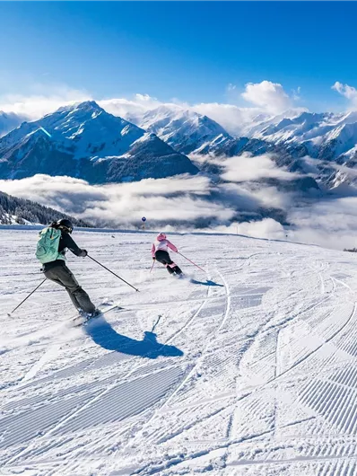 Two skiers are skiing down a snow-covered slope. In the background, there are majestic mountains and a clear blue sky.