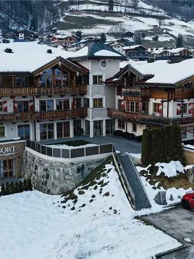 A picturesque hotel in alpine style, surrounded by snow-covered mountains. There are several cars in the parking lot and a quiet, inviting atmosphere.