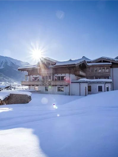 A charming house in a snow-covered landscape with mountains in the background. The sun shines bright, giving the scene a golden glow.