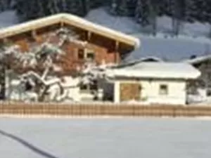 Eine verschneite Landschaft mit traditionellen Häusern und Bäumen. Im Hintergrund erheben sich bewaldete Berge unter einem klaren blauen Himmel.