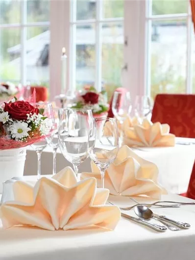 Een elegante restauranttafel met een witte tafellaken en kunstzinnig gevouwen servetten. Rode stoelen en bloemenarrangementen zorgen voor een feestelijke sfeer.
