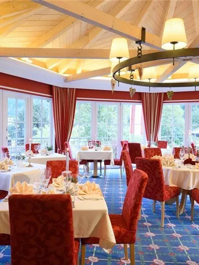 Een elegant restaurant met rode stoelen en tafellakens. Grote ramen laten veel licht binnen en bieden uitzicht naar buiten.