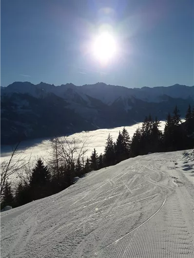 Een schilderachtig winterlandschap met verse sneeuw en bergen op de achtergrond. De zon schijnt boven een wolkendek dat de vallei bedekt.