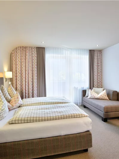 Ein modernes Hotelzimmer mit zwei Einzelbetten und einem Sofa. Helle Farben und viel Tageslicht schaffen eine einladende Atmosphäre.