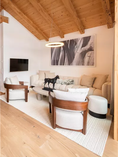 Ein modernes Wohnzimmer mit einer hellen Couch, Sesseln und einem Holztisch. Die Decke hat sichtbare Holzbalken und ein großes Kunstwerk hängt an der Wand.