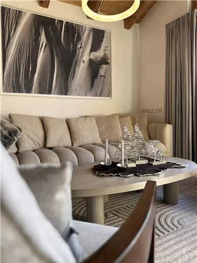 Ein elegantes Wohnzimmer mit einer hellen Couch und einem runden Tisch. An der Wand hängt ein großes Kunstwerk, und der Raum hat große Fenster mit Vorhängen.