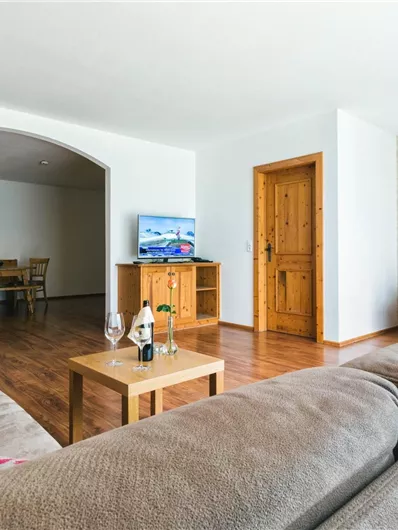 Ein helles Wohnzimmer mit gemütlichen Sofas und einem Holztisch. An der Wand hängt ein Bild, und es gibt dekorative Elemente wie Blumen und Kissen.