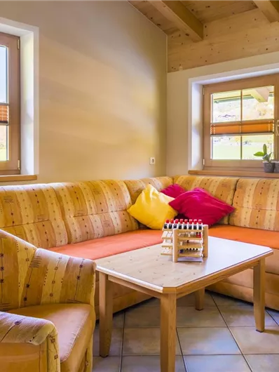 Een gezellige zithoek met een comfortabele bank en een tafel. Door de ramen stroomt zonlicht en er zijn enkele planten te zien.