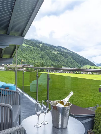 Een balkon met uitzicht op een groene weide en bergen op de achtergrond. Er zijn comfortabele meubels en een champagnesfles met glazen op de tafel.