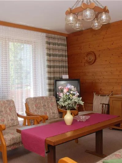 Ein gemütliches Wohnzimmer mit Holzverkleidung und einem großen Fenster. Die Einrichtung besteht aus einem Tisch, zwei Sesseln und einer Kommode.