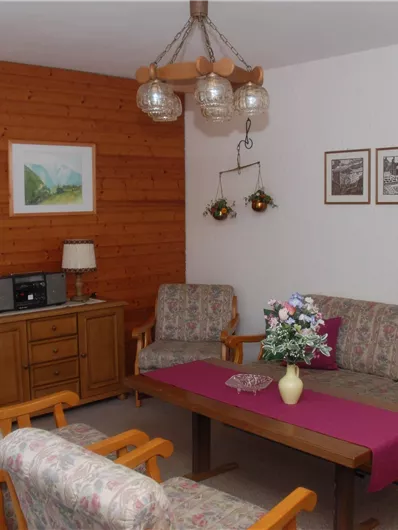 Ein gemütliches Wohnzimmer mit Holzwänden und einem einfachen Sofa. Eine Blumenvase steht auf dem Tisch, während Bilder an der Wand hängen.