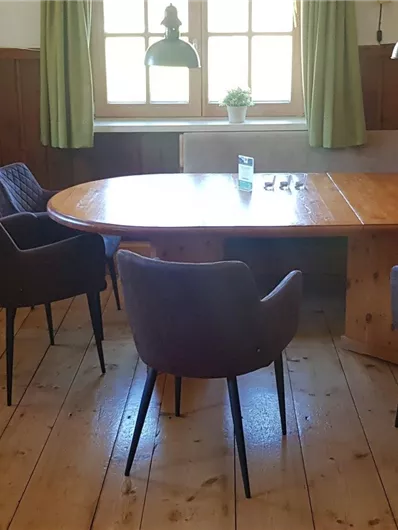 Ein gemütlicher Raum mit einem großen Holztisch und schwarzen Stühlen. Die Wände sind aus Holz, und es gibt grüne Vorhänge sowie eine Fensterbank mit Pflanzen.