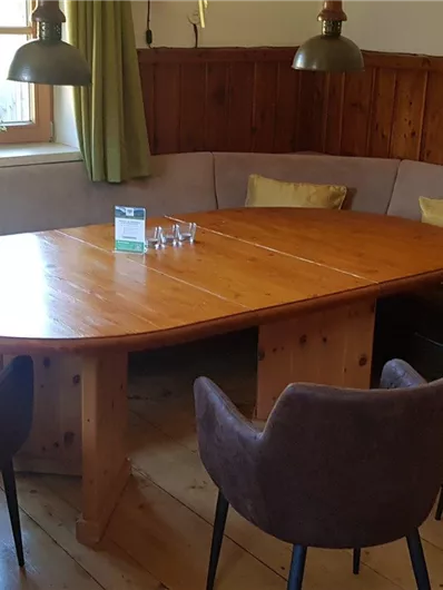 Ein gemütlicher Raum mit einem runden Holztisch und bequemen Stühlen. Große Fenster lassen viel Tageslicht herein und die Wände sind teilweise aus Holz.