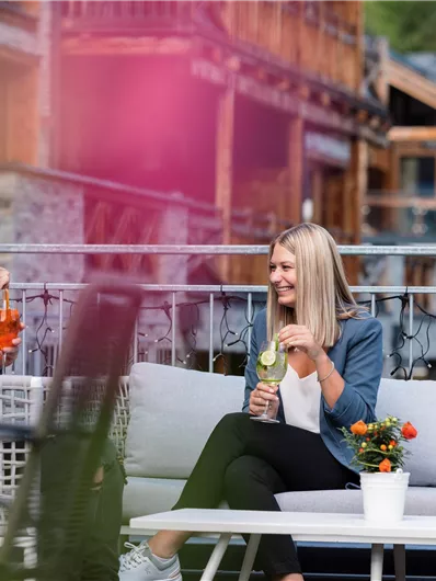 A young woman is sitting on a couch and enjoying a drink. In the background, wooden houses and flowers can be seen.
