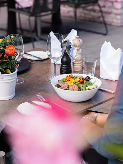A table outdoors with a colorful salad bowl and a flower in a pot. In the background, there are more tables and guests visible.