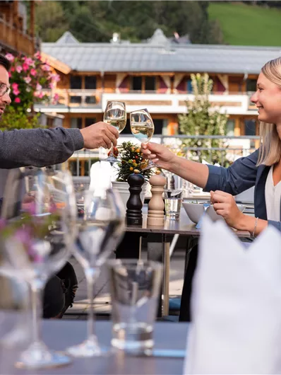 A couple toasts with glasses and enjoys a meal outdoors. In the background, beautiful buildings and green spaces can be seen.