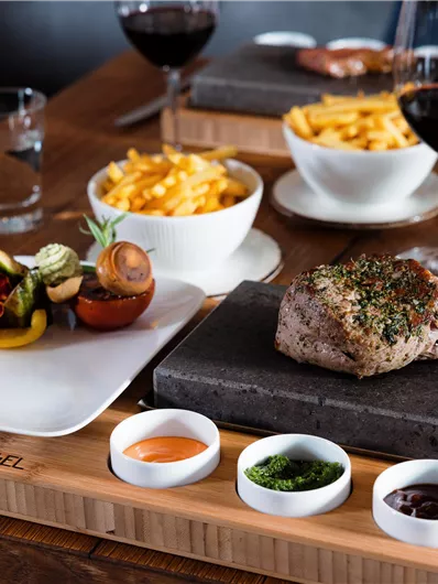 A delicious steak on a hot stone, accompanied by sauces and vegetables. Served with French fries and red wine in the background.