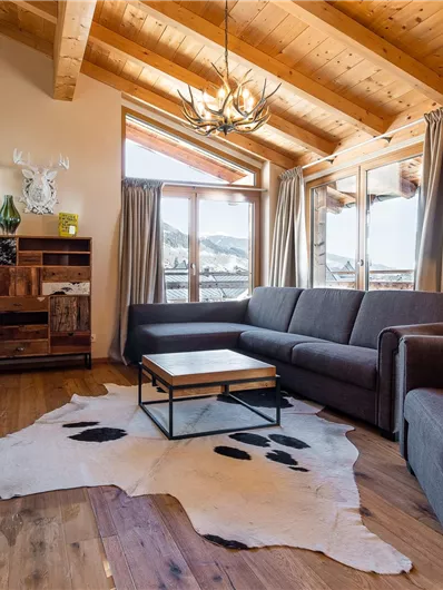 A cozy living room with a modern sofa and a wooden table. A fireplace creates a warm atmosphere and large windows provide a view outside.