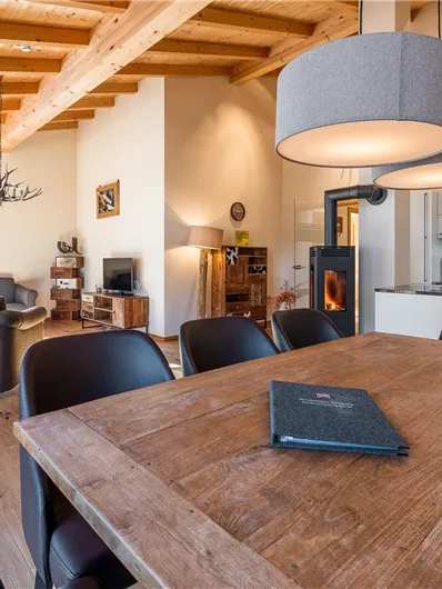 A modern living room with wooden ceilings and generous windows. A wooden dining table is the centerpiece, surrounded by black chairs.