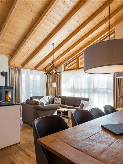 A modern living and dining room with a wooden beam ceiling. Large windows let in plenty of light and create a cozy atmosphere.