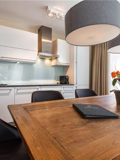 A modern kitchen with a large wooden table and elegant chairs. Stylish lamps hang above the table, and a plant adds a fresh accent to the space.