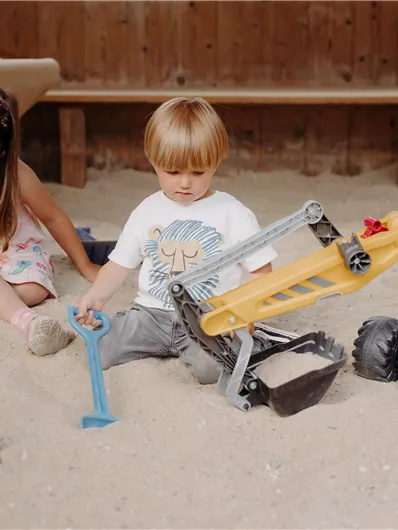 Zwei Kinder spielen im Sandkasten. Ein Junge mit einer gelben Baufahrzeug und ein Mädchen mit einer Schaufel sind beschäftigt.
