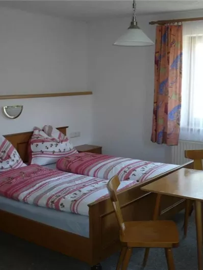 Ein einfaches Schlafzimmer mit einem Doppelbett und bunten Kissen. Ein Tisch mit Stühlen und ein großes Fenster sorgen für Helligkeit.