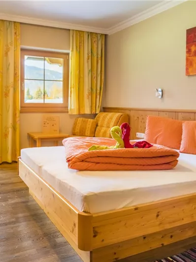 Een gezellige slaapkamer met een tweepersoonsbed en oranje kussens. Grote ramen laten veel daglicht binnen en bieden uitzicht naar buiten.