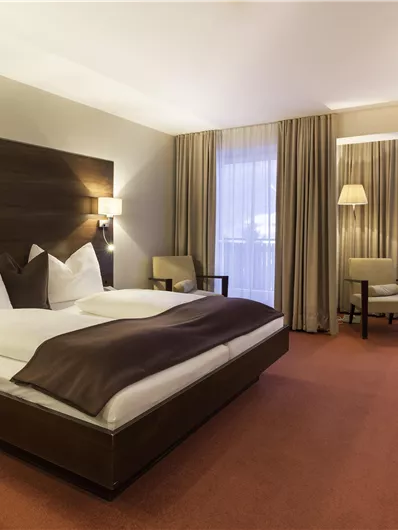 A modern hotel room with a double bed and stylish pillows. The decor is cozy and bright with windows that let in daylight.