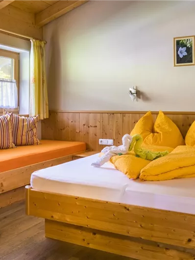 Een gezellige slaapkamer met een tweepersoonsbed en een beklede zithoek. Grote ramen laten veel daglicht binnen en de houten wandbekleding zorgt voor een warme sfeer.