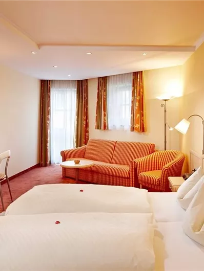 Een gezellig hotelkamer met twee bedden, een rode bank en een bureau. Er zijn grote ramen en vriendelijke, warme kleuren.