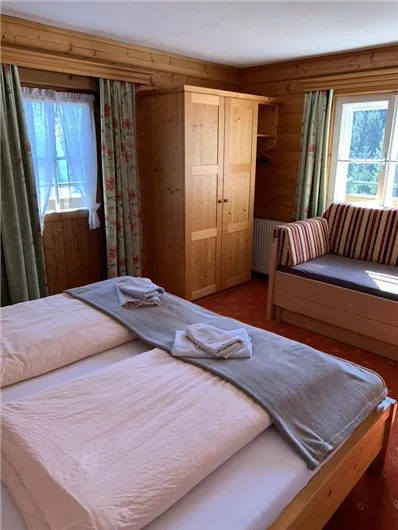 Ein gemütliches Schlafzimmer mit Holzverkleidung, zwei Betten und einem klassischen Kleiderschrank. Fenster mit Vorhängen lassen natürliches Licht herein, und eine Sitzecke lädt zum Entspannen ein.